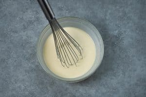 A glass bowl holding smooth pale yellow batter being mixed with a wire whisk.
