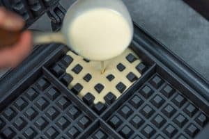 A metal ladle pouring smooth pale yellow batter onto the lower grid of a black square waffle maker.