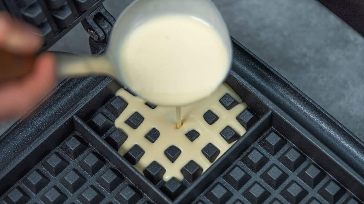 A metal ladle pouring smooth pale yellow batter onto the lower grid of a black square waffle maker.