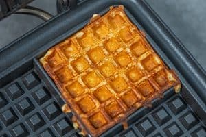 A freshly cooked square golden brown waffle resting on the black lower grid of an open waffle maker.