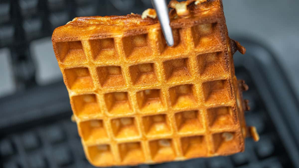 Metal tongs holding up a freshly cooked square golden brown waffle.